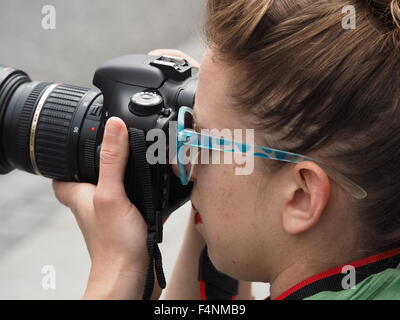 Fotografo femmina indossando occhiali da sole guardando attraverso il mirino della fotocamera reflex digitale con obiettivo zoom Foto Stock