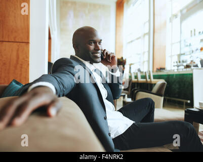 Felice giovane imprenditore rilassante seduta sul divano a hotel lobby e fare una chiamata telefonica in attesa di qualcuno. Foto Stock