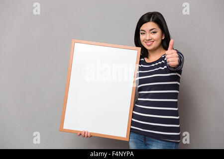 Ritratto di una donna sorridente di contenimento della scheda in bianco su sfondo grigio e mostrando il pollice in alto Foto Stock