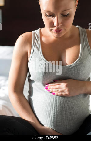 Donna incinta si siede sul letto con la sua mano a pancia Foto Stock