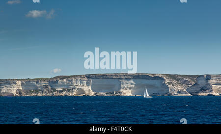 Un bianco barca a vela sul blu del mare Mediterraneo insieme contro le bianche scogliere vicino a Bonifacio in Corsica del sud con i cieli blu abov Foto Stock