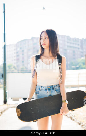 Ginocchio la figura del giovane bello asiatico marrone lungo i capelli dritti donna skater tenendo uno skateboard, ponendo all'aperto in città, affacciato sulla destra - sportivi, pensieroso concept Foto Stock