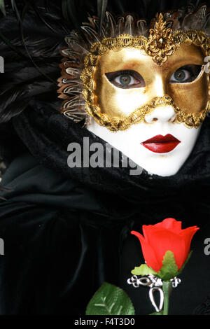 Maschera dell'amore: Haunting donna con incredibili occhi in golden feathered maschera con pizzo nero abbigliamento e una rosa rossa al Carnevale di Venezia Foto Stock