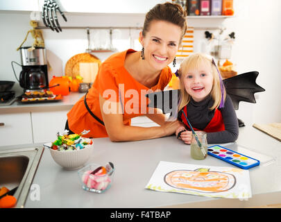 Sorridente ragazza bionda in costume bat con la giovane madre creazione di decorazioni di halloween con carta e vernici. Preparazione per un partito è in pieno svolgimento. Tradizionale autunno holiday Foto Stock