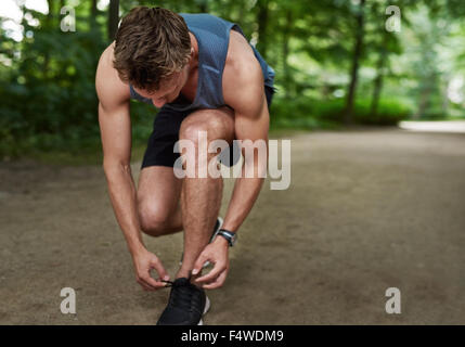 Montare muscolare pareggiatore maschio la piegatura verso il basso la sua legatura lacci delle scarpe in una via attraverso un lussureggiante parco alberato in uno stile di vita sano concetto Foto Stock