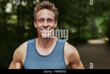 Sorridente muscolare bello giovane uomo in piedi all'aperto in un parco alberato in sportswear guardando la telecamera, testa e spalle p Foto Stock