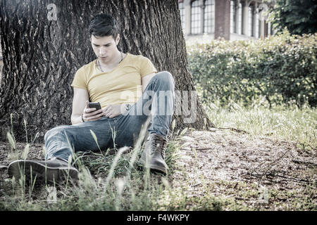 Attraente giovane nel parco in appoggio contro la struttura, utilizzando il cellulare di tipo messaggio, rilassato in una soleggiata giornata estiva Foto Stock