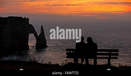 I turisti seduti su un banco di lavoro guardando a Porte d'Aval, arco naturale in Chalk Scogliere di Etretat al tramonto, Alta Normandia, Francia Foto Stock