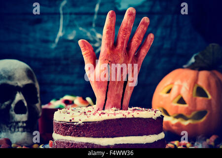 Una torta rossa del velluto e rabboccato con una sanguinosa mano in una scena spaventoso per la festa di Halloween con una zucca intagliata, un cranio e ragnatele Foto Stock