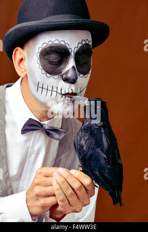 Ritratto di un uomo messicano con calaveras trucco, indossando il filtro bow tie e cappello bowler, baciando un corvo nero Foto Stock