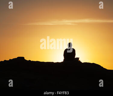 Silhouette di un uomo solitario seduto di fronte al tramonto della linea costiera della California. Foto Stock