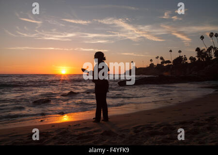 Silhouette di una donna in piedi con un telefono di fronte al tramonto della linea costiera della California. Foto Stock