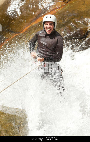 Un uomo che indossa una muta nera e un casco bianco sta scivolando lungo una cascata rocciosa, tenendosi su una corda. Sorride in generale con un'espressione di eccitazione mentre l'acqua gli spruzza intorno. La scena Foto Stock