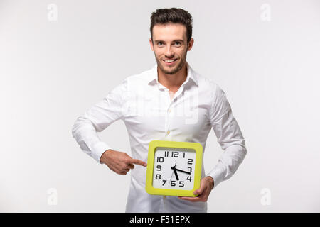 Ritratto di un imprenditore sorridente in camicia puntare il dito su orologio da parete isolata su uno sfondo bianco Foto Stock