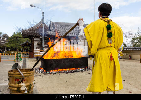 Giappone, Nishinomiya, Mondo Yakujin tempio buddista. Sacerdote in giallo Yamabushi accappatoi, in piedi a guardare il fuoco consuma l'anno di buona fortuna fascino. Foto Stock