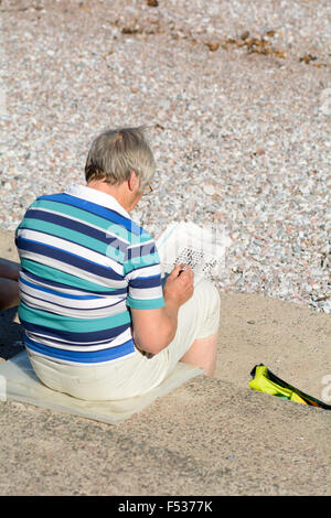 Uomo seduto sui gradini a Oddicombe beach facendo un cruciverba di giornale sul soleggiato settembre giornata in Torquay, Devon, Inghilterra Foto Stock