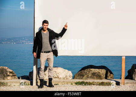 Bel giovane uomo in piedi accanto al grande vuoto tabellone bianco, display o bordo, outdoor dalla riva del lago Foto Stock