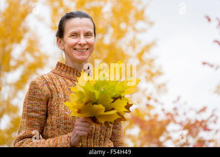 Un sorridente adulto donna caucasica è raccolta giallo foglie di acero in autunno nel parco Foto Stock
