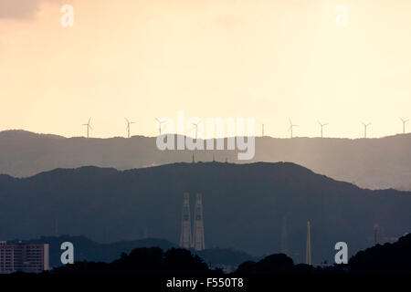 Wakayama, Giappone, stagliano le turbine eoliche sulla cima delle montagne in lontananza con vari camini industriali in primo piano. Tempesta di pioggia, ma alcuni la luce del sole. Foto Stock