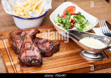 Costolette di agnello grigliate su un tagliere di legno con una porzione di insalata e patatine fritte Foto Stock