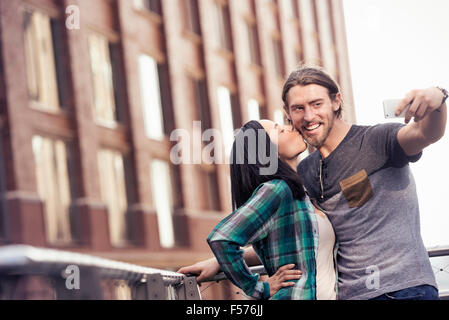 Una donna baciare un uomo sulla guancia in posa per una selfie da un grande edificio della città Foto Stock