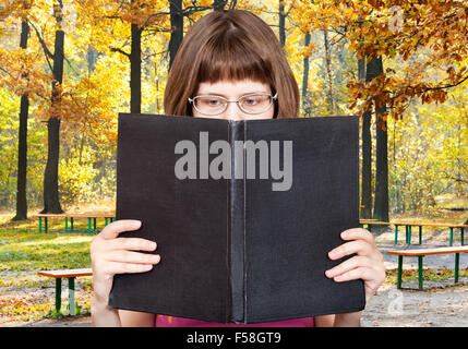 Ragazza con gli occhiali legge big book con coperchio bianco e giallo autunno foresta sullo sfondo Foto Stock