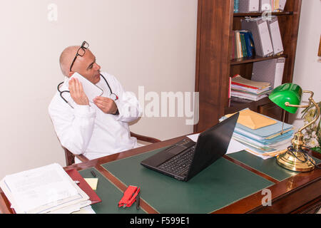 Sconcertato medico nel suo studio, utilizza una tavoletta davanti al suo computer portatile. Utilizzare le nuove tecnologie. Nel suo studio professionale, egli è sitti Foto Stock
