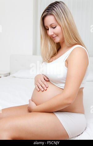 Donna incinta seduta sul letto e guardando sul suo ventre Foto Stock