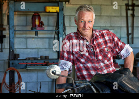 Uomo di mezza età accanto alla sua vecchia bici del motore. Il suo esterno resistente viene ammorbidito mediante i suoi occhi blu e consolante sorriso. Foto Stock