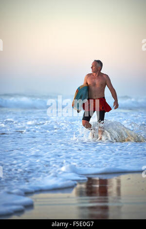 Uomo di mezza età a piedi in onda oceano lavare in possesso di una tavola da surf. Soft Early Morning Light lambisce la deliziosa scena. Foto Stock