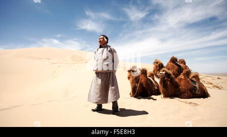 Un cammello mongolo herder si erge con orgoglio davanti ai suoi cammelli nel bel mezzo delle dune di sabbia del deserto del Gobi. Mongolia Foto Stock