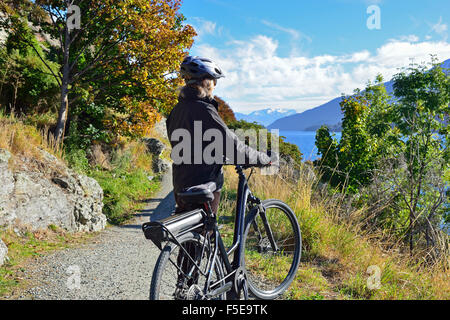 Signora ciclista su una bicicletta elettrica E-Bike, guardando il panorama sulle rive del Lago Wanaka, Frankton, Nr Queenstown, Isola del Sud, Nuova Zelanda Foto Stock