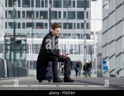 Ritratto di un allegro giovane uomo seduto sulla valigia alla stazione guardando al telefono cellulare Foto Stock