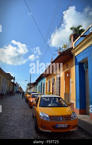 Giallo taxi parcheggiato in una riga in una strada di ciottoli con case colorate in Trinidad, Cuba Foto Stock