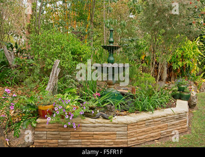 Giardino decorativo caratteristica con un basso muro di pietra, fontana ornata, piante in contenitori e orlate da prato, fiori, arbusti, alberi Foto Stock