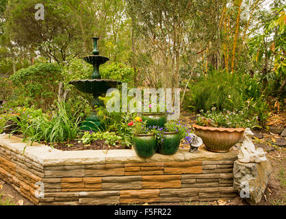 Giardino decorativo caratteristica con un basso muro di pietra, fontana ornata & piante in contenitori, orlata da rigogliosi arbusti, bambù, alti alberi Foto Stock