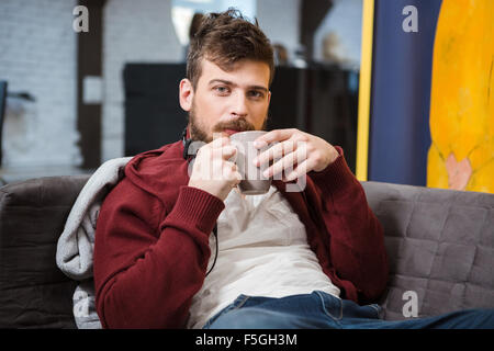 Rilassati bello ragazzo seduto sul divano grigio e di bere il caffè Foto Stock