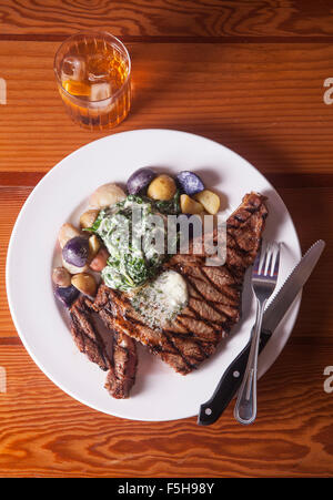 Vista aerea della bistecca con patate lesse e spinaci coltello e forchetta e cocktail sulla tavola di legno Foto Stock