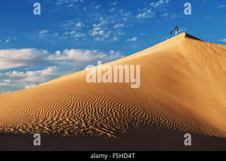 Un tradizionale vestito uomo marocchino sorge su una duna di sabbia contro un nuvoloso cielo blu. Foto Stock