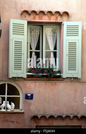 Esterno di un rosa cottage francese con un pallido persiane verdi su una finestra con tende di pizzo Foto Stock