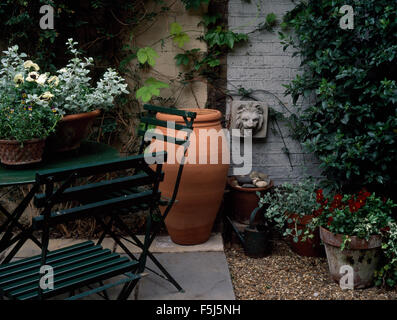 Foglie di piante in vaso sul piccolo tavolo con sedie a doghe su una città patio con un alto vaso in terracotta e una lapide in pietra Foto Stock