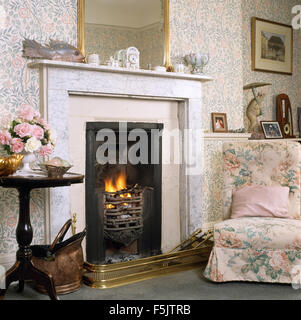 Sedia floreali accanto al caminetto con fuoco illuminato in salotto con carta da parati floreale Foto Stock