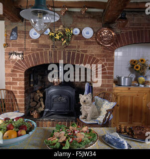 West Highland terrier seduti su una sedia accanto al camino con una stufa a legna esposta una parete di mattoni in un cottage cucina Foto Stock