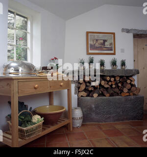 Argento servire piatto e cesto di verdure su una tavola di pino in un cottage cucina con i registri ordinatamente impilati nel camino Foto Stock