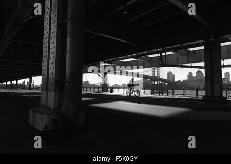 Un uomo di cicli lungo la East River Esplanade, New York, sotto il Franklin Roosevelt East River Drive cavalcavia come il sole che sorge Foto Stock