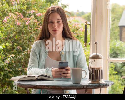 Ritratto di giovane donna seduta a tavola, tenendo lo smartphone Foto Stock