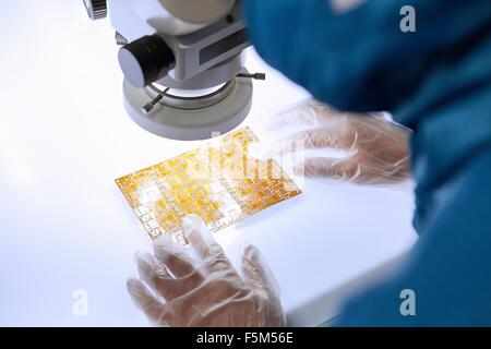 Al di sopra della spalla vista del lavoratore di sesso femminile che usando microscopio per esaminare il circuito flessibile in elettronica flessibile fabbrica camera pulita Foto Stock