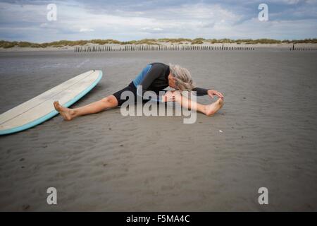 Senior donna seduta sulla sabbia, stretching, tavola da surf accanto a lei Foto Stock