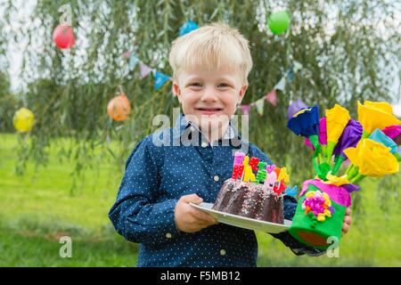 Ritratto di ragazzo portando la torta di compleanno e fiori di carta in giardino Foto Stock