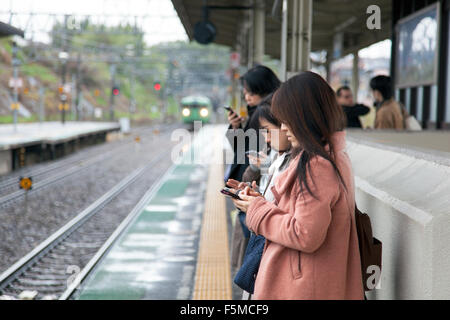 Ragazze utilizzando i telefoni cellulari in attesa di un treno, Giappone Foto Stock